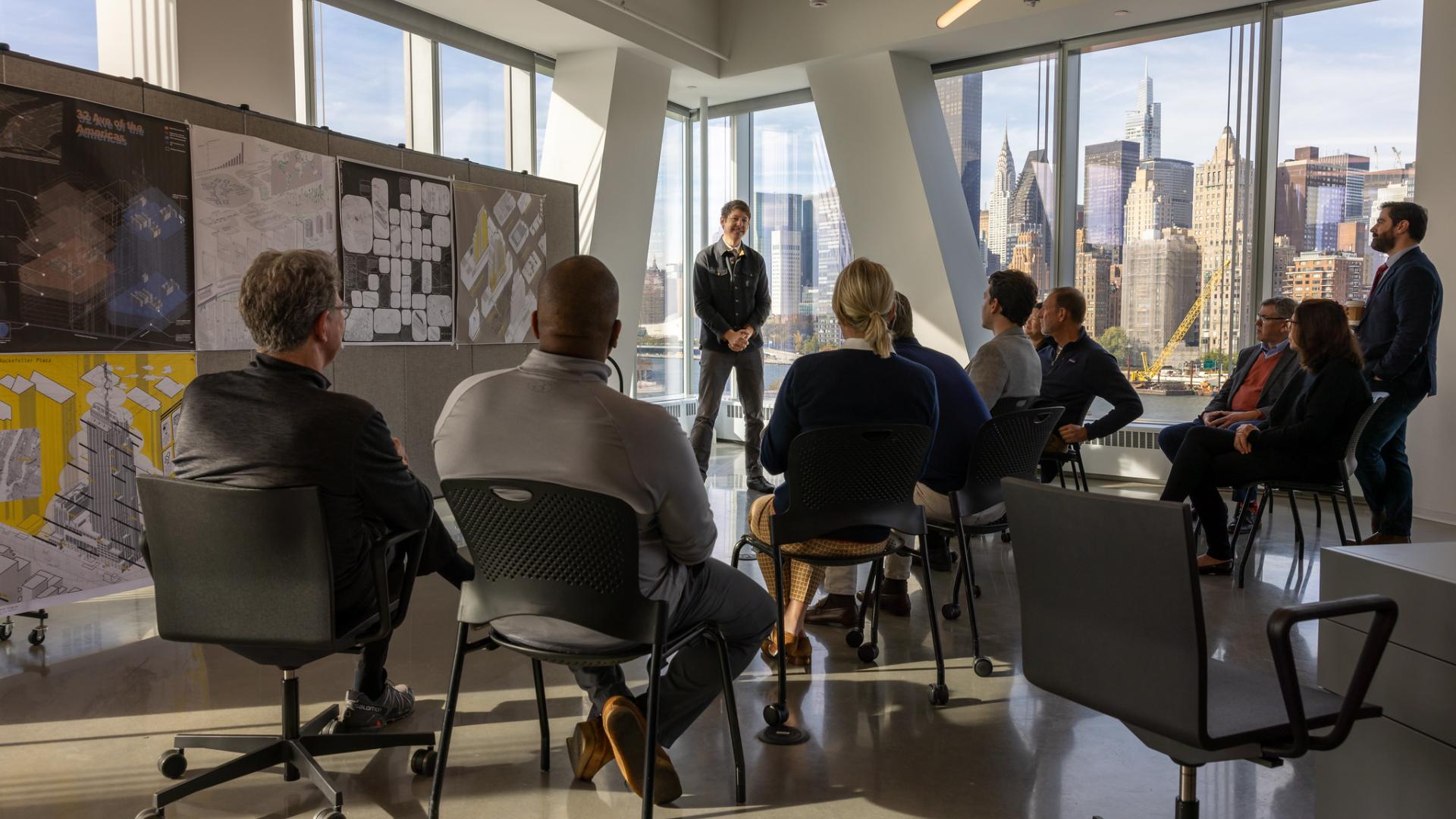 A group of people listen to someone give a presentation, standing in front of architectural drawings.