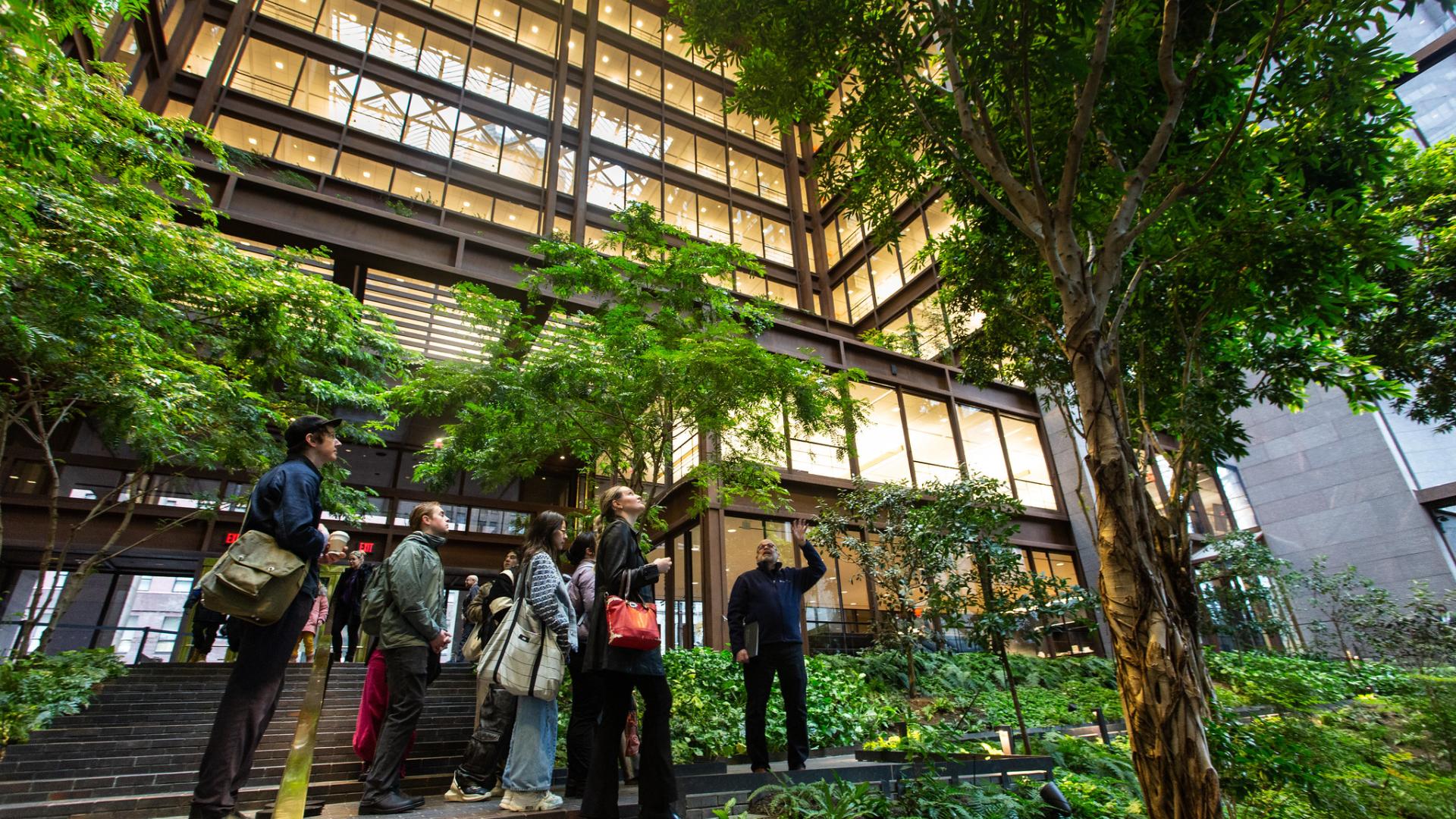 A group of people listen to someone speak outside of a building among trees and greenery.