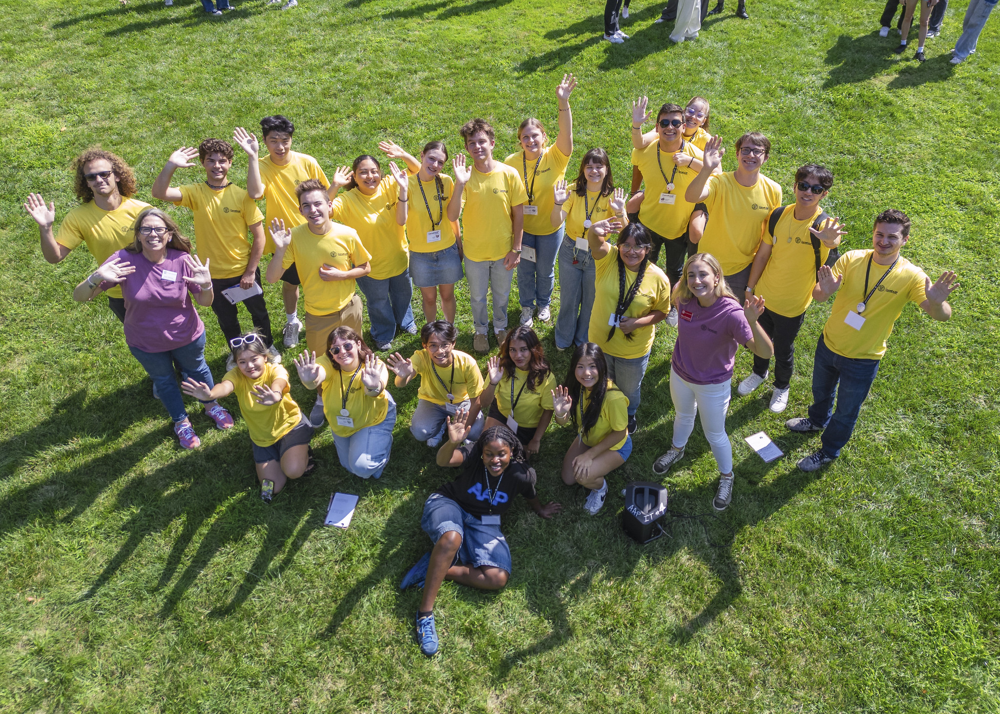 A group of people dressed in yellow and pink shirts stand outside and look up at the camera and wave.