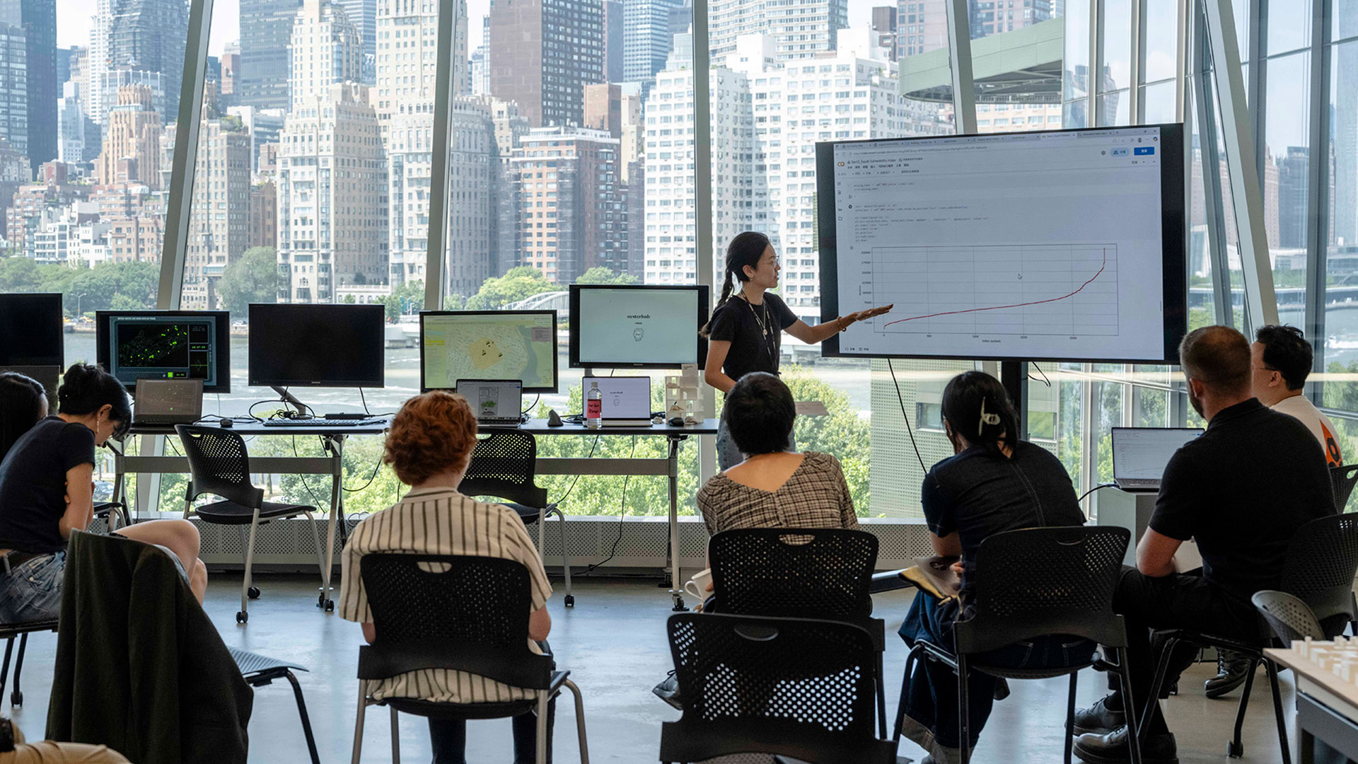 A person presents data on a screen while others sit and listen.