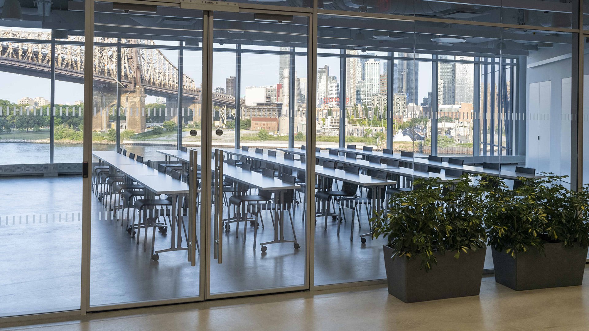 A view outside of a glass walled room with many tables and chairs inside, and a distant view of New York City in the background.