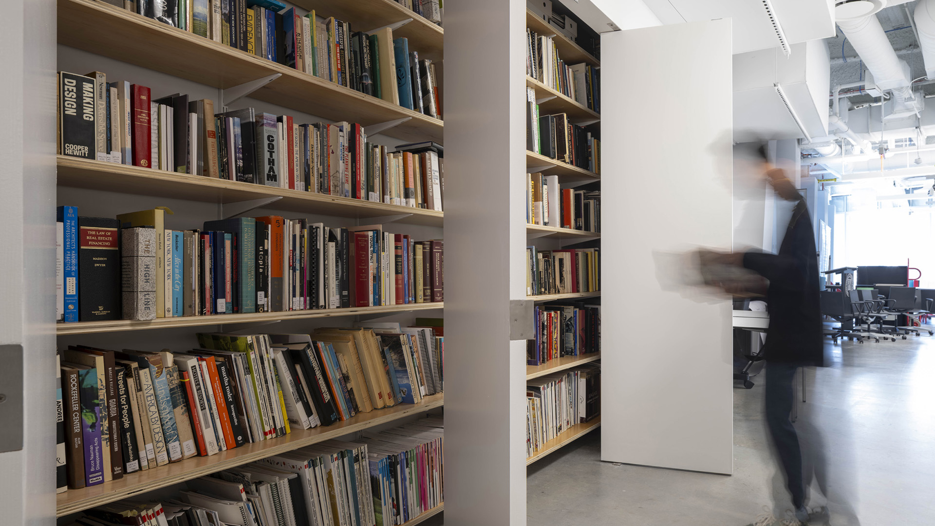 A person browses a book in a library in front of a wall of books.