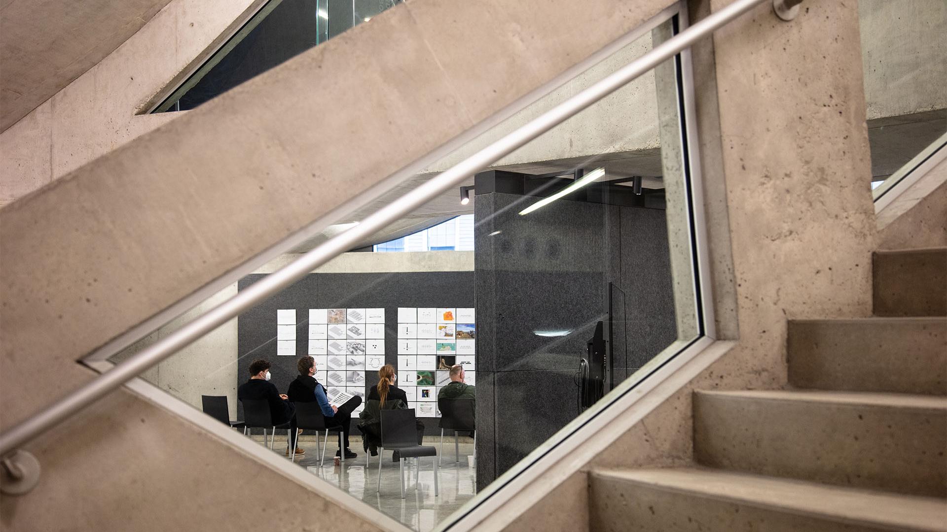 View from the staircase of a wall that is covered in architectural drawings, with people sitting in chairs listening to a presentation.
