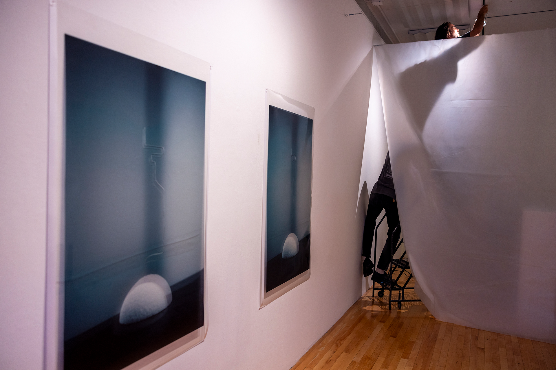 A person adjusts ceiling lights on a ladder, with prints hanging on the wall.