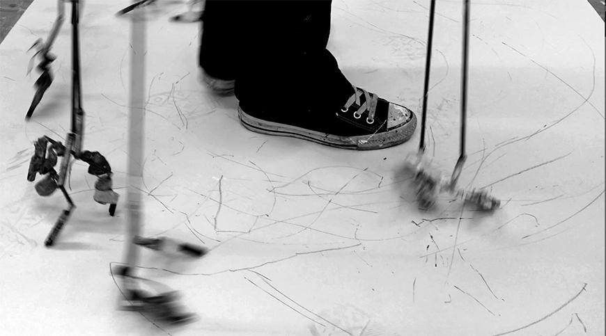 A person wearing sneakers is standing amidst long sticks that are touching the ground and making marks on the floor.