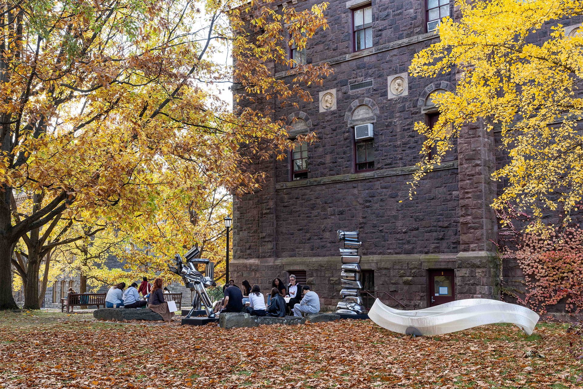 Exterior view of a stone building with autumn trees and people sitting among sculptures in a circle.