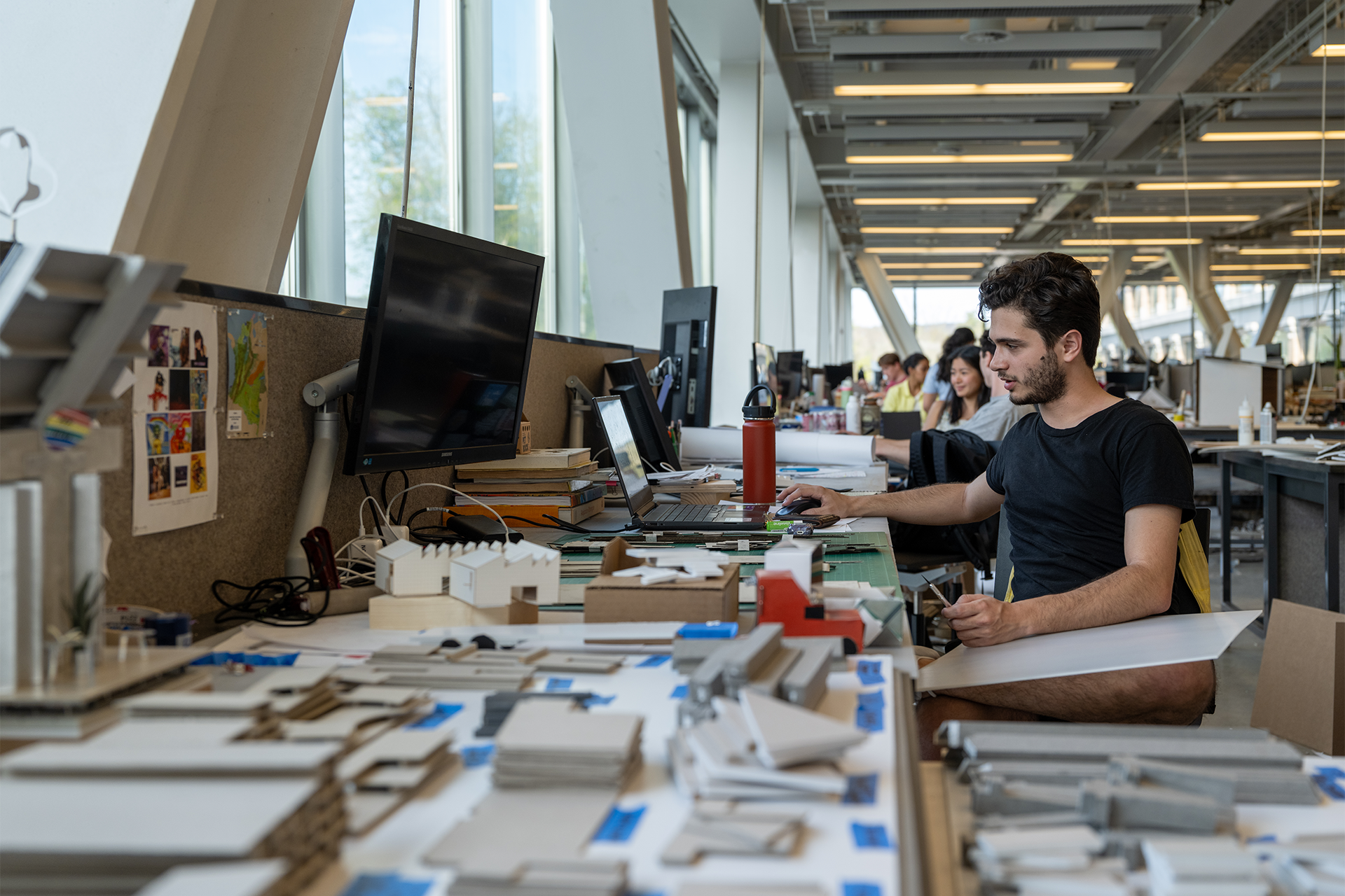 View of students sitting at their studio desks that are covered with architectural models and components.