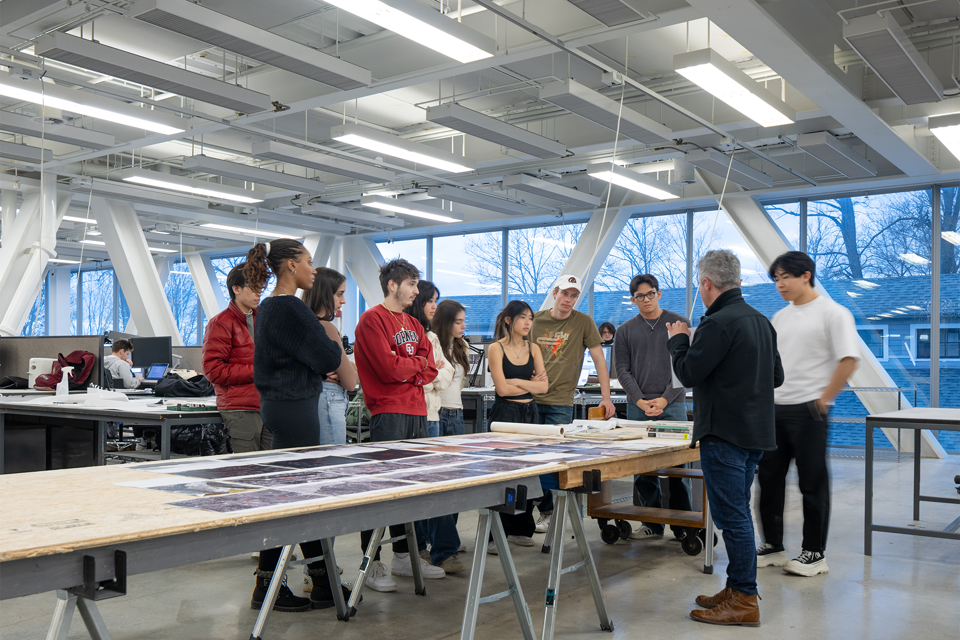 A group of students listens to someone present papers on a table.