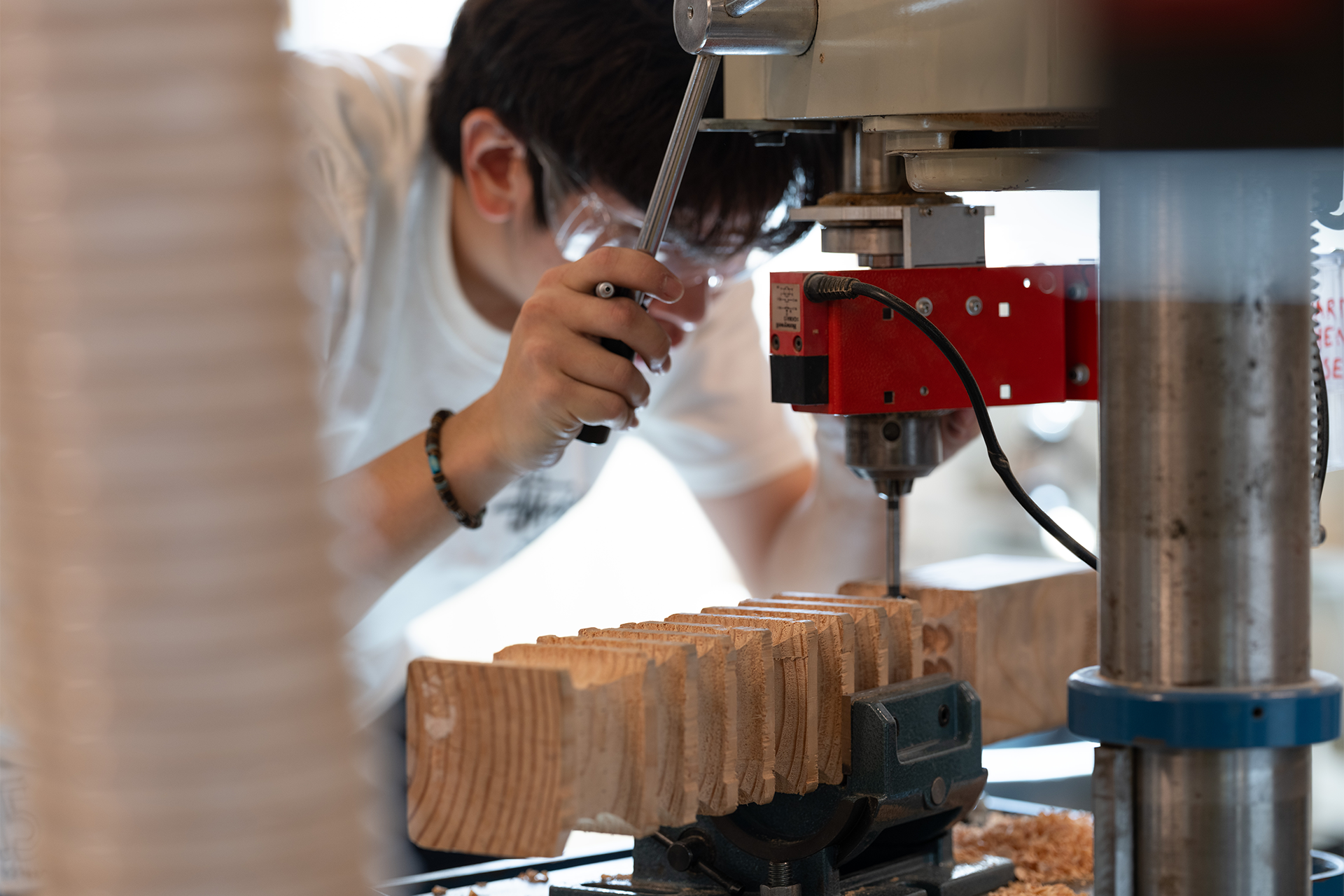 A person uses a drill press tool to shape a long rectangular block of wood.