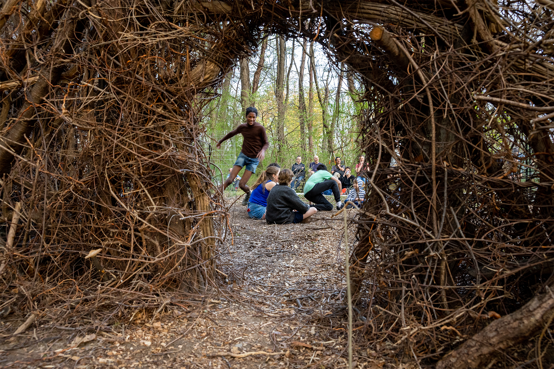 A group of people play outside, with the camera person positioned behind an archway of brambles and sticks.