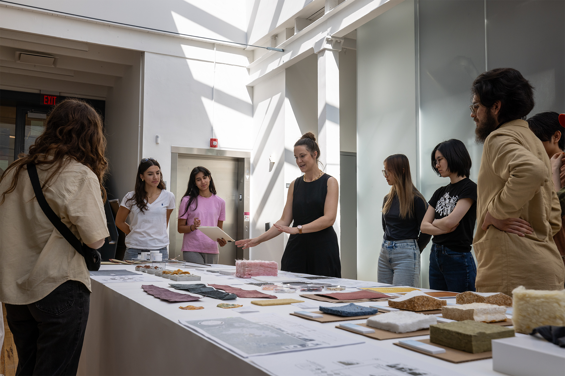 A person speaks to a group of people around a long table that is covered with different samples of materials.