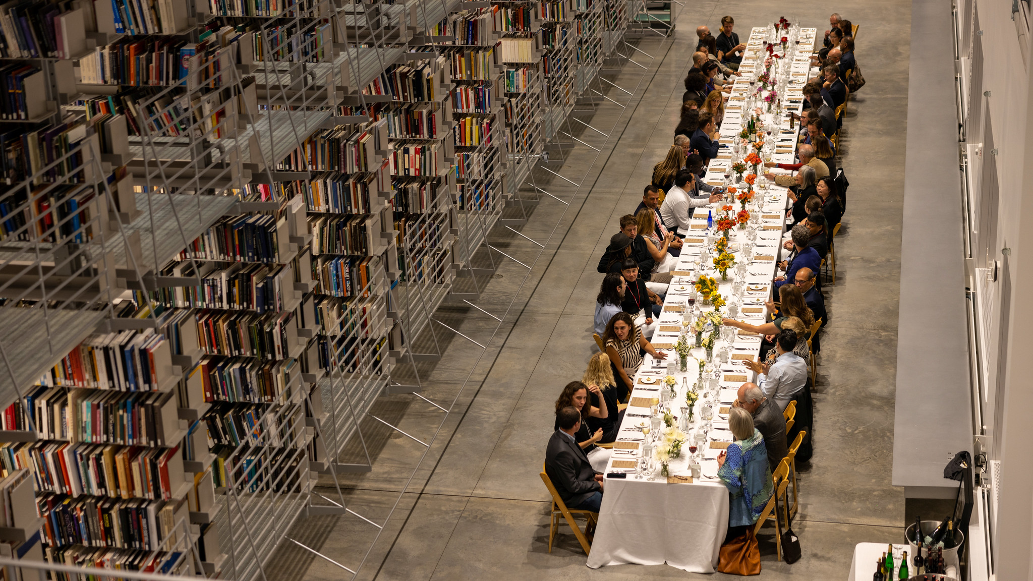 An overhead photo of a long dining table with people sitting on either side, with library book stacks on the side.