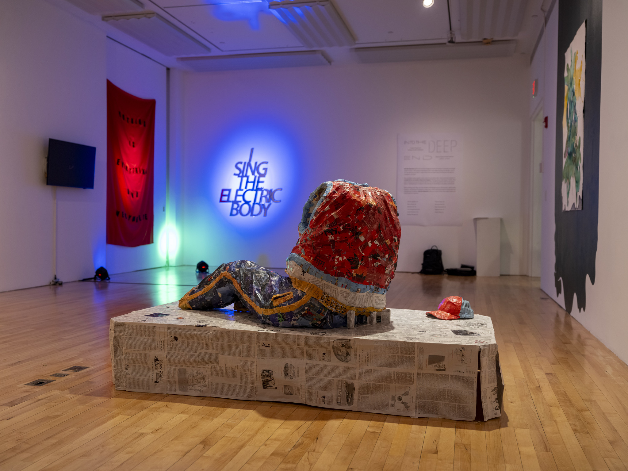 A raised platform covered in newspaper with a sculpture on top. In the background, there is a red cloth panel hanging from the wall, and a neon sign on the wall next to it.