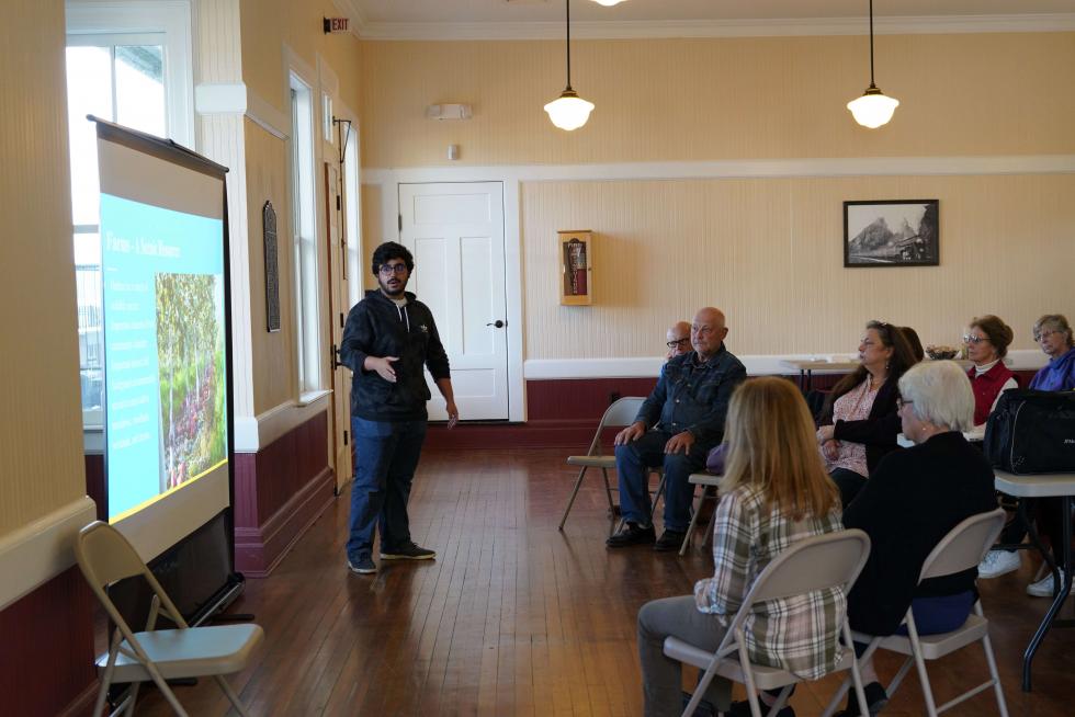 Person gives a presentation at the front of a room while an audience sits in metal chairs watching the presentation.
