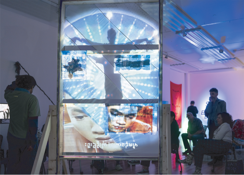 A film is projected on the side of a wooden framed canvas, and people are seated in the background.