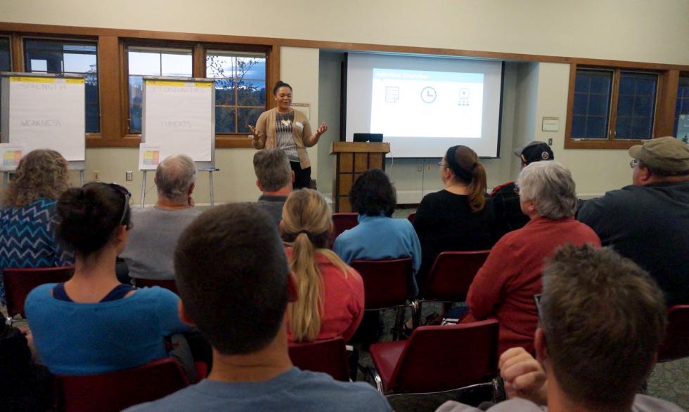 A person gives a presentation at the front of a classroom while other people sit in red chairs and listen.