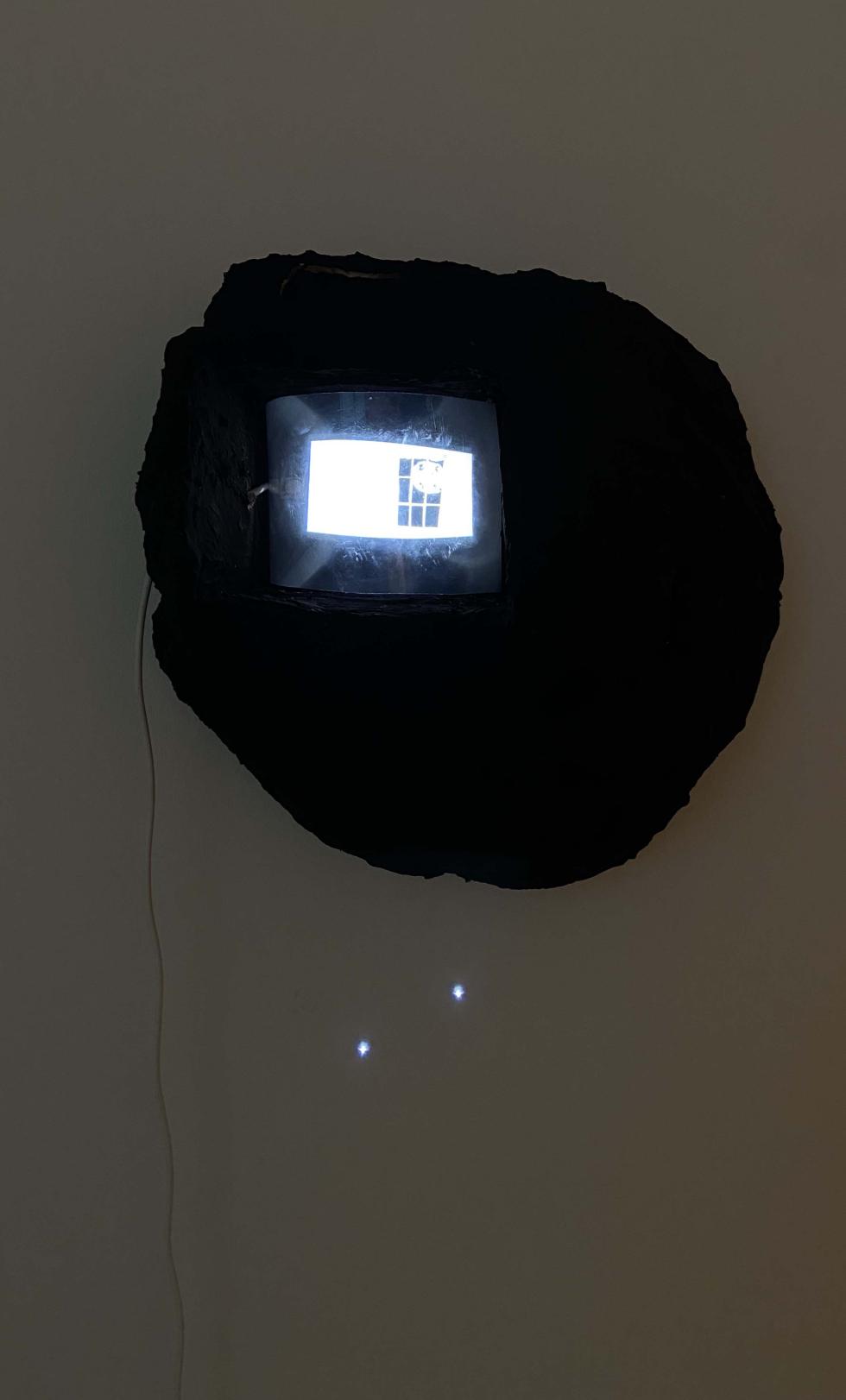 A black sculpture hangs on a gallery wall with a small projection inside it.