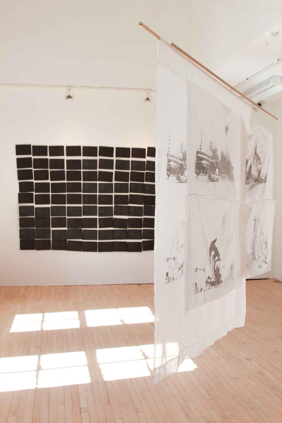 Installation view of two artworks. A grid of black paper squares on one wall, and two hanging panels of white fabric.