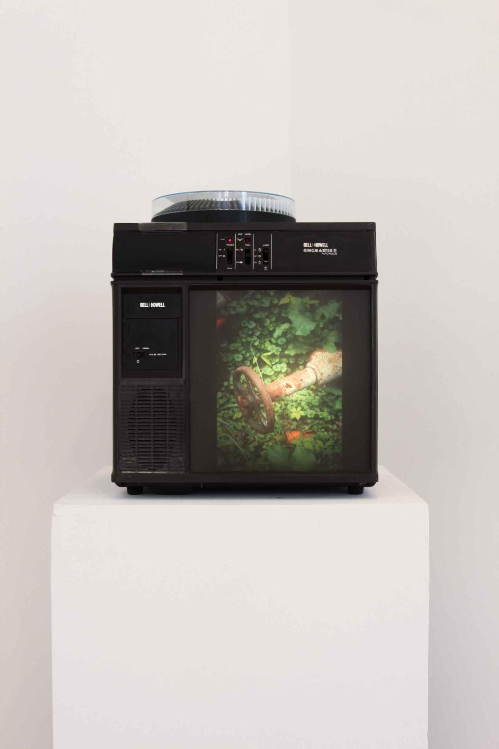 A slide projector sits on a white pedestal and displays an image of a water spigot.
