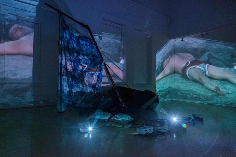 A darkened room with video equipment on the ground and two projections of a person lying on the ground.