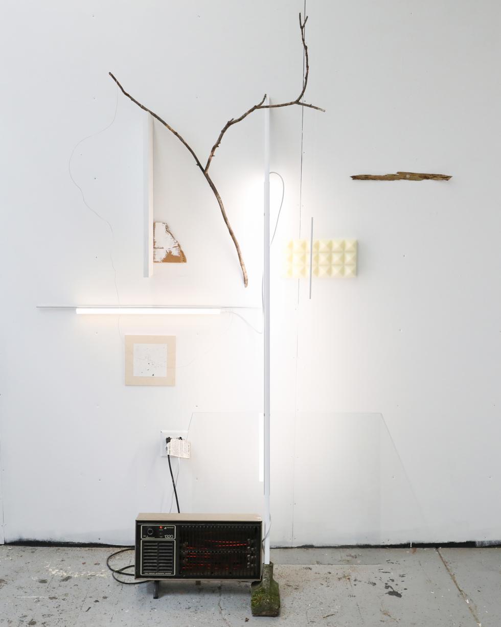 Linear lights on a wall illuminate a sculpture of wood, a radio, and paper.