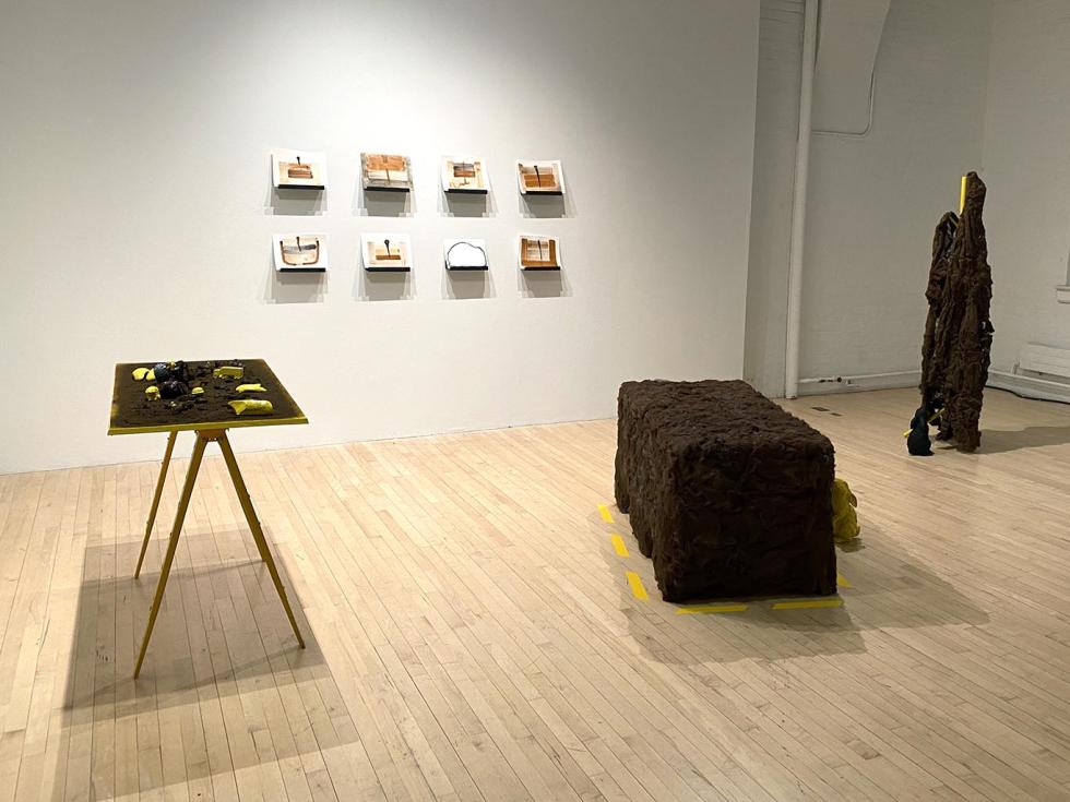 Gallery view of an exhibition that features a large bench, brown sculpture, a table with objects on top, and eight small shelves with paintings.