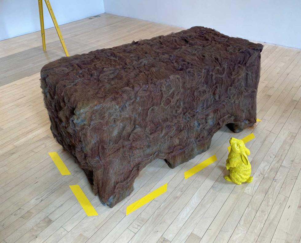 A large brown rectangular sculpture with a small yellow bunny sculpture looking at it.
