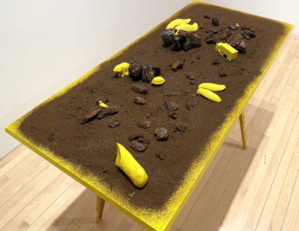 A yellow table with dirt on top of it, scattered with found objects and porcelain casts of objects.