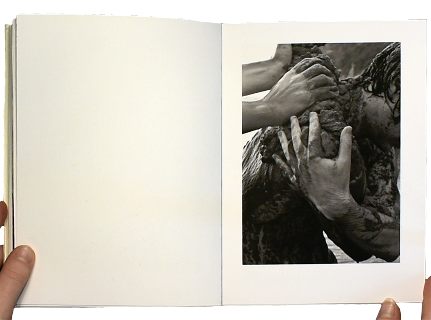 A person opens a book to a page with a photograph of a person covering another person in mud.