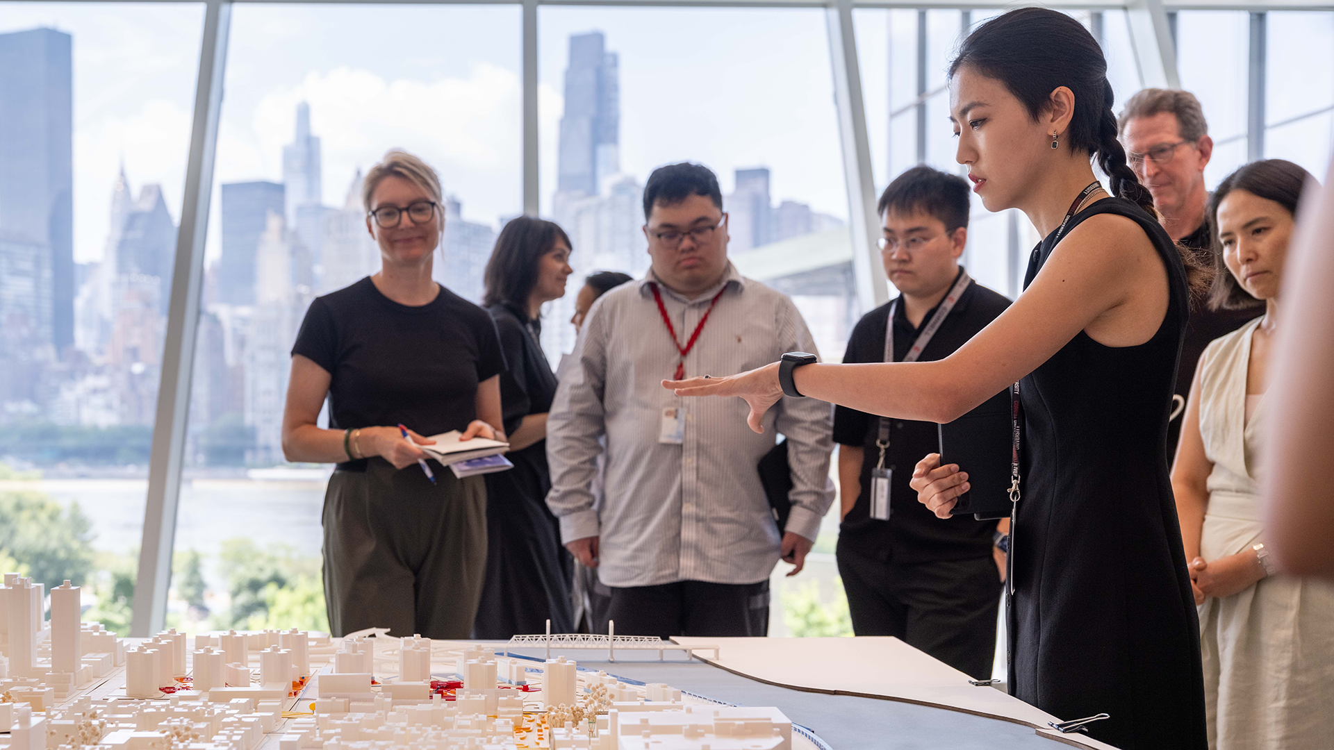 A person presents an architectural model on a table while other people watch them.