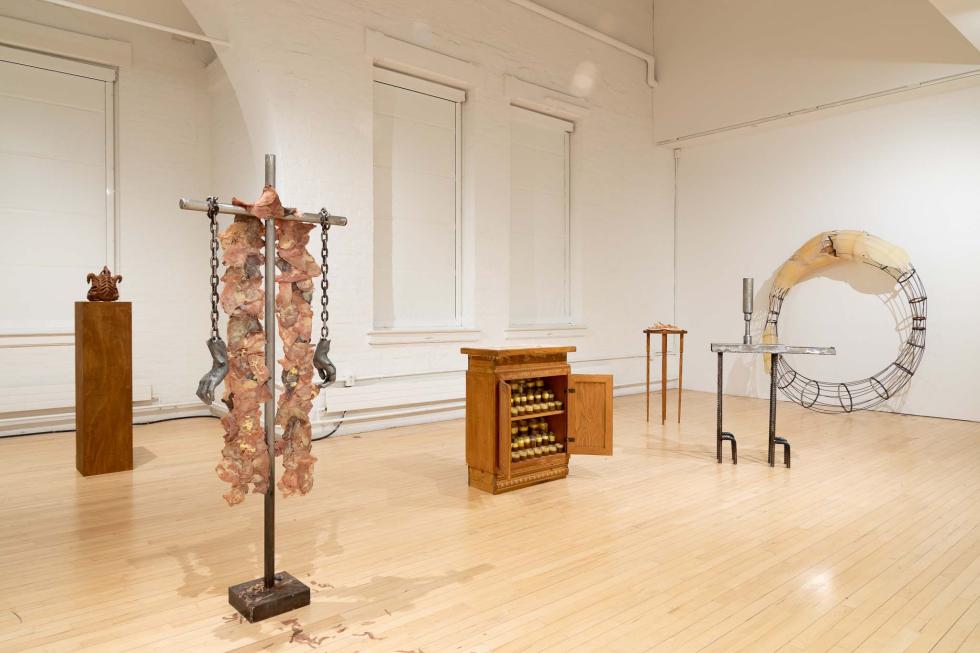 Gallery view of an exhibition that includes metal sculptures, a wooden cabinet filled with small objects, and a wooden pedestal and table.