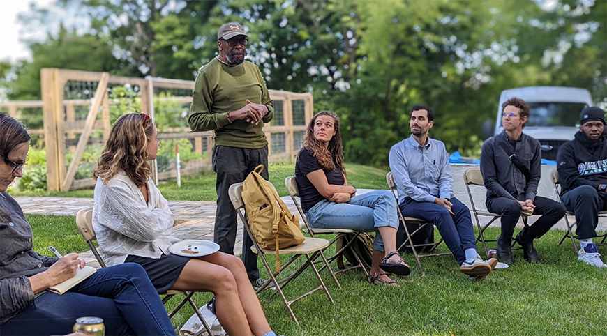 People sit in a circle of chairs outside and one person stands to talk to the group.