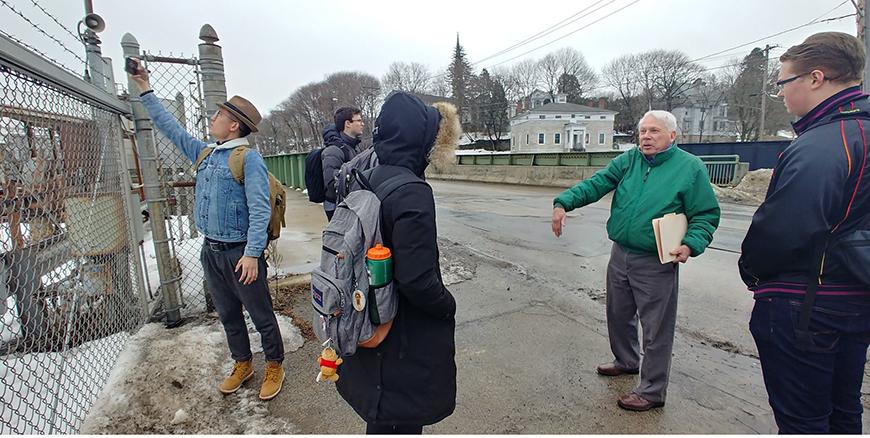 People gather in a circle on the side of a paved road next to a wire gate.