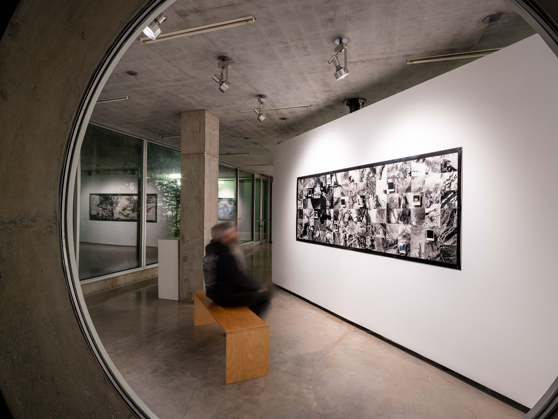 A person sits on a gallery bench and views a long print of aerial photographs.