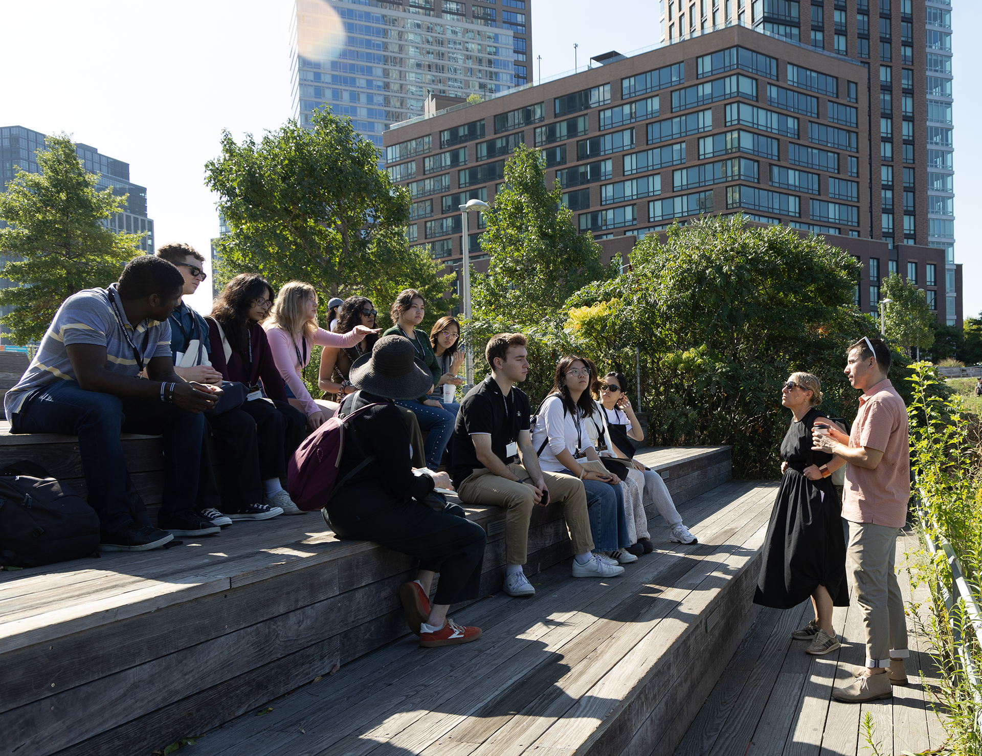 People sit on an amphitheater in an park setting while two people present to them. There are large buildings in the background.
