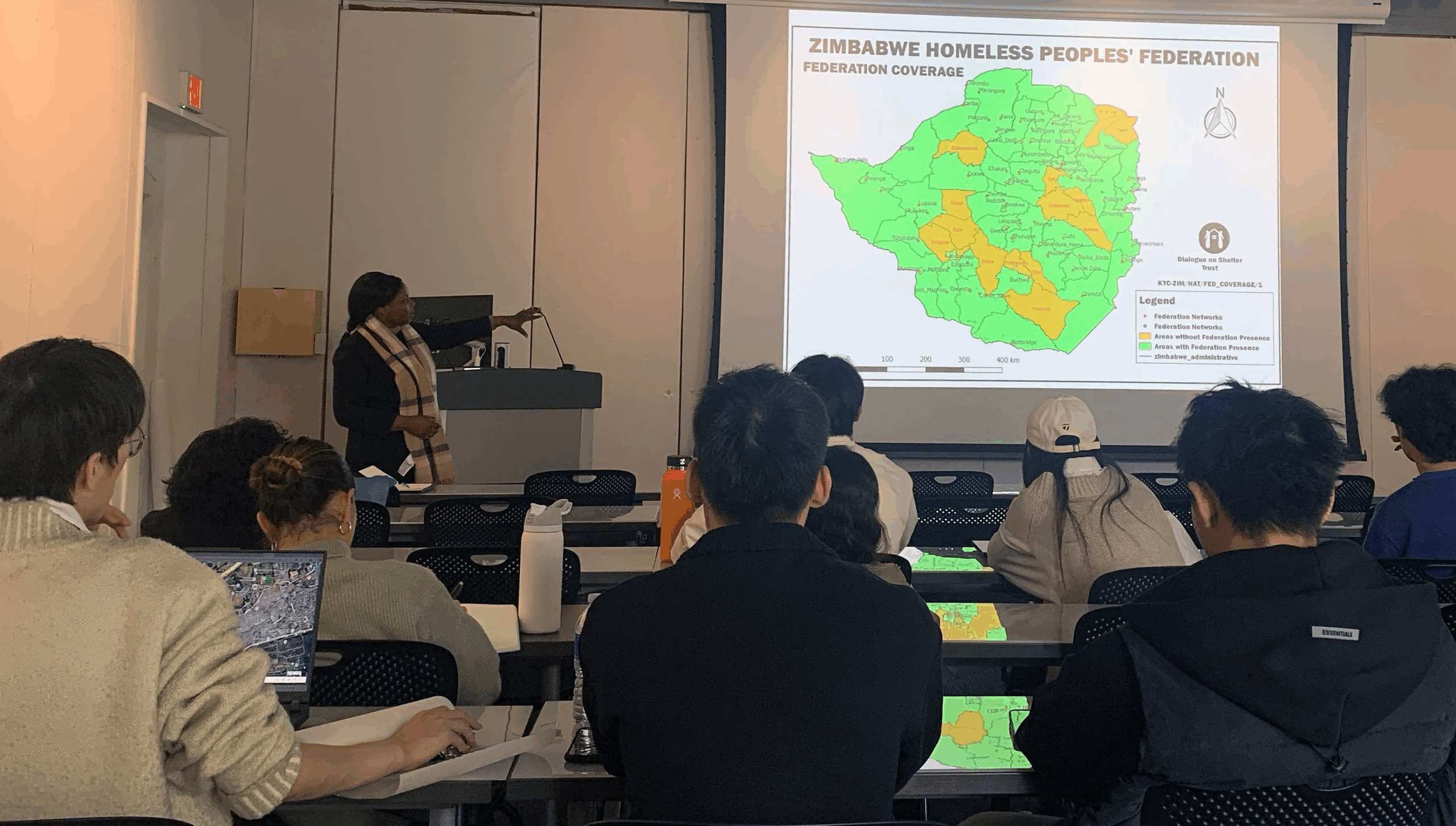 Classroom presentation with people in rows in the audience watching one person pointing to a presentation of a map on a projector.