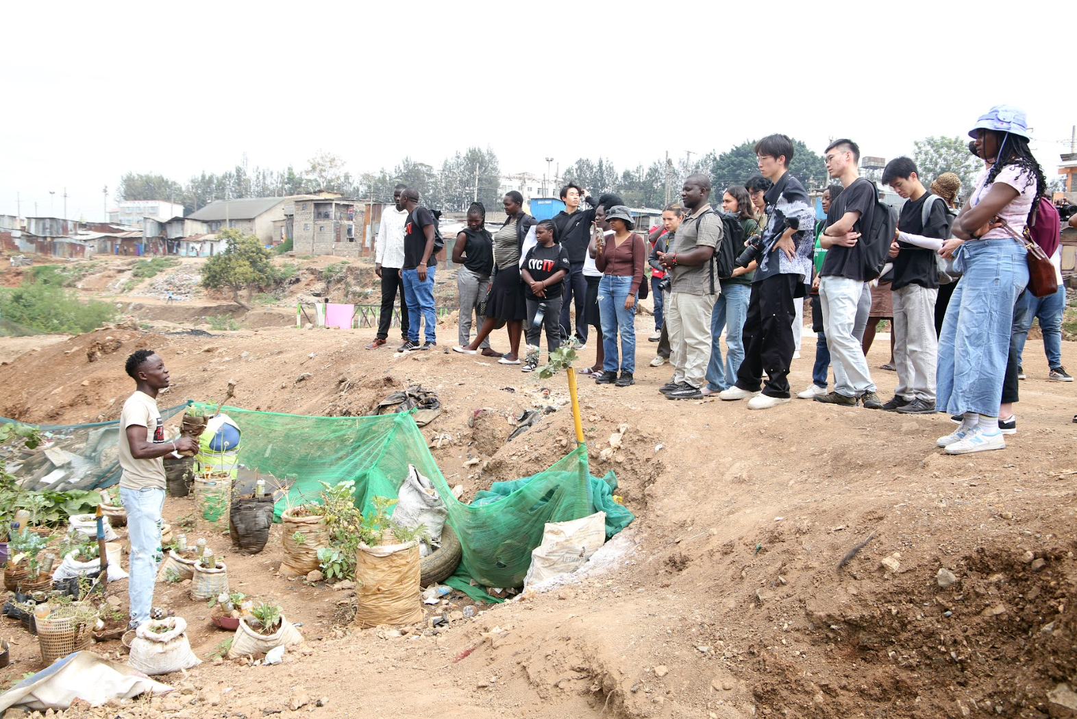 A group of people watch another person in a small valley with plants inside of a variety of bags.