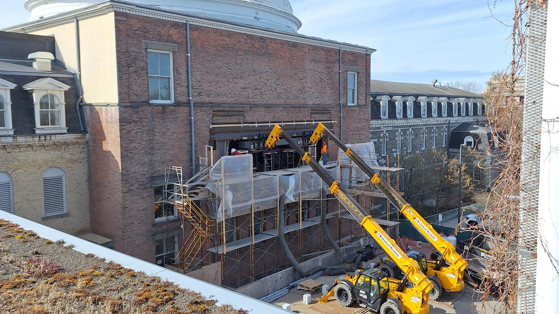 Exterior of a brick building with two cranes delivering materials to to people on scaffolds.