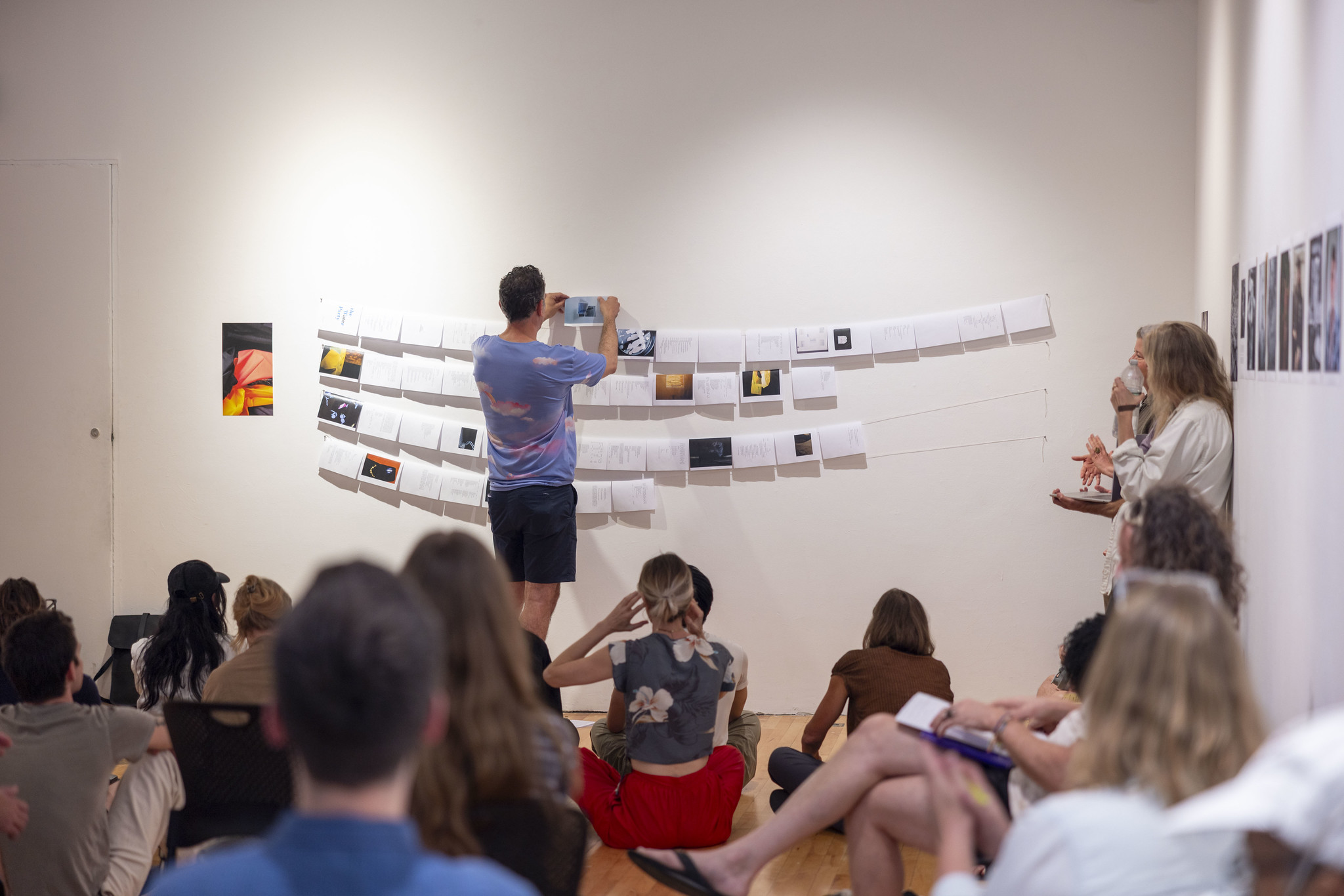 Gallery with people sitting on the ground watching a person pin up artwork to the wall.