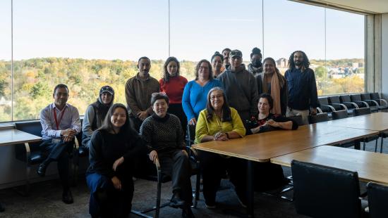 A group of people pose together in a conference room with large windows overlooking an autumn landscape, seated and standing around long tables.
