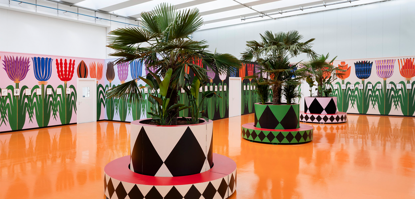 Interior of an exhibition with cylindrical planters covered in checkered black, green, and red designs, with an orange floor and walls that have 2d flowers painted on them.
