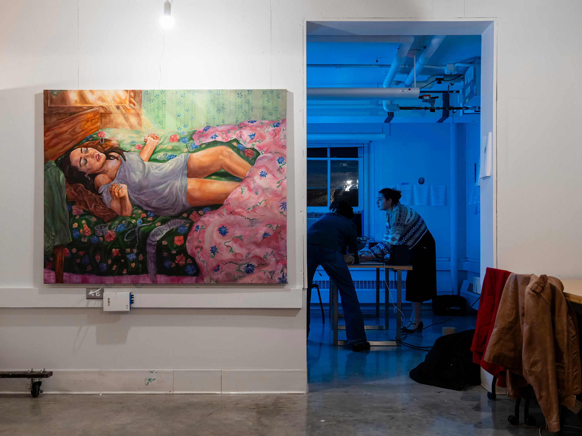 Interior of an art studio with a large painting of a person on a bed. The art studio leads into a common space with two people using a projector at a table.