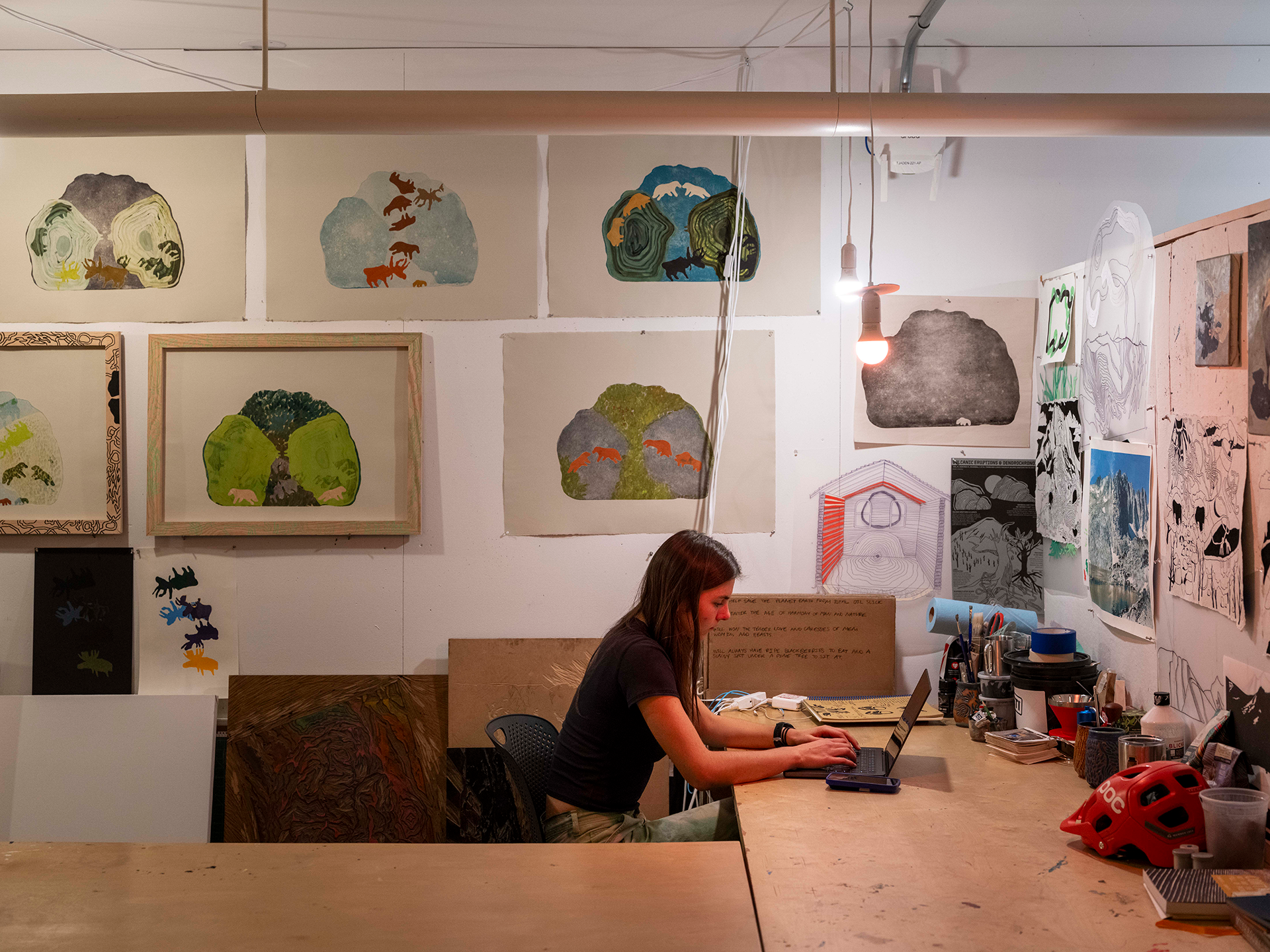 Interior of a student's art studio with artwork lining the walls, and a student sitting at a desk on a laptop.