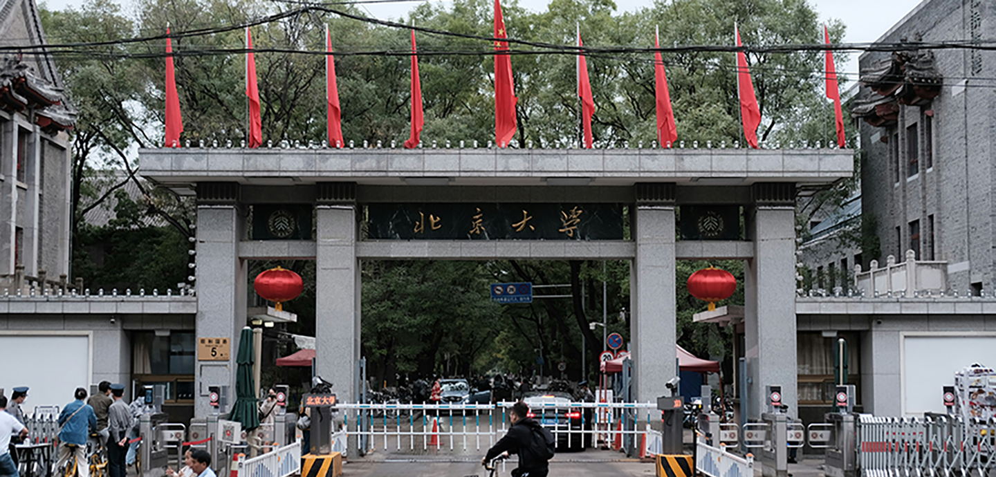 A grand stone entrance gate decorated with red flags and lanterns leads into a busy tree-lined campus.