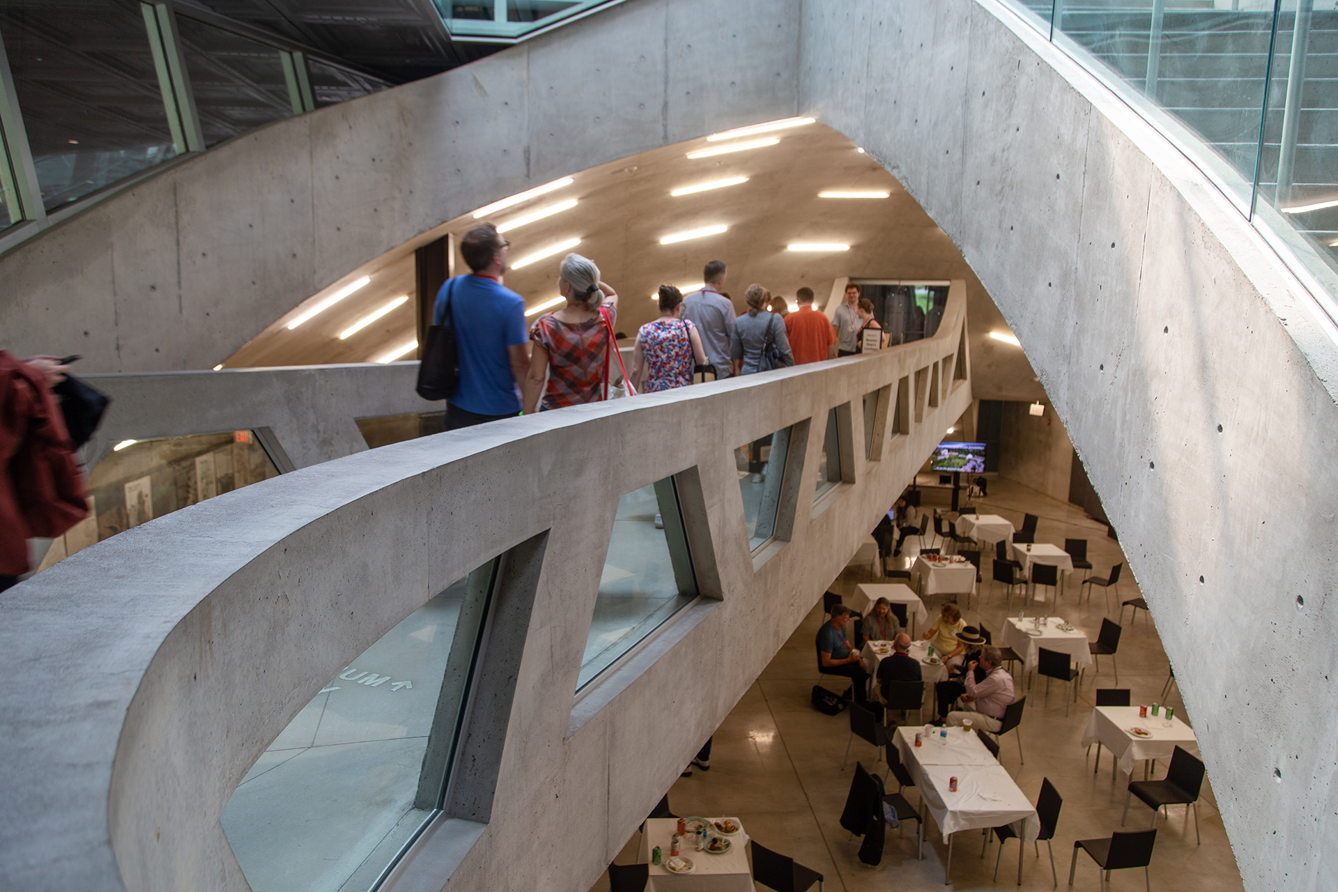 Concrete walkway bridge leading to an auditorium. People line the walkway. Below the walkway is a series of tables with people sitting at each table.