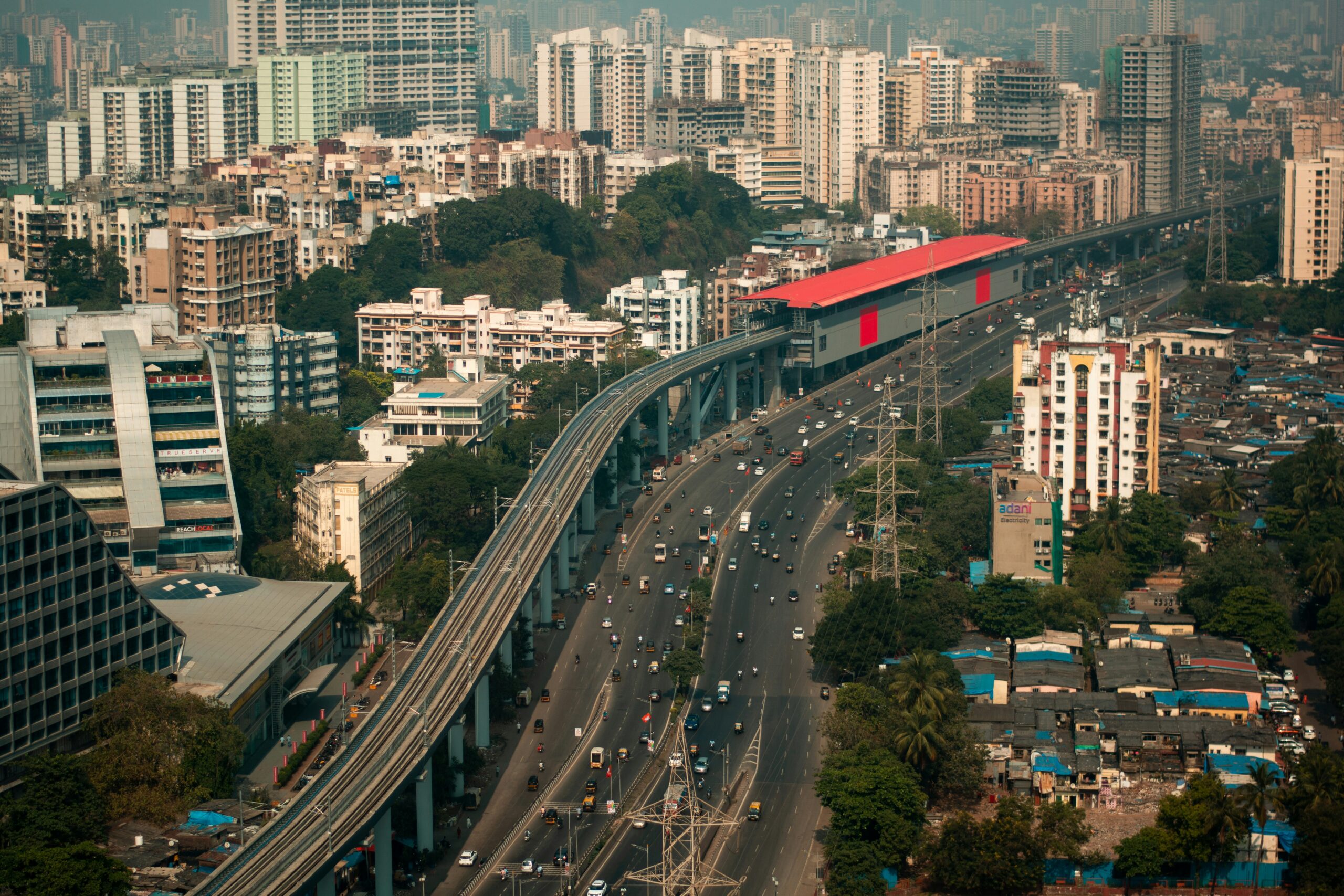 Aerial view of a busy multilane highway and elevated metro line curving through dense Mumbai high-rises, with a red-roof transit station (metro) and tightly packed neighborhoods below.
