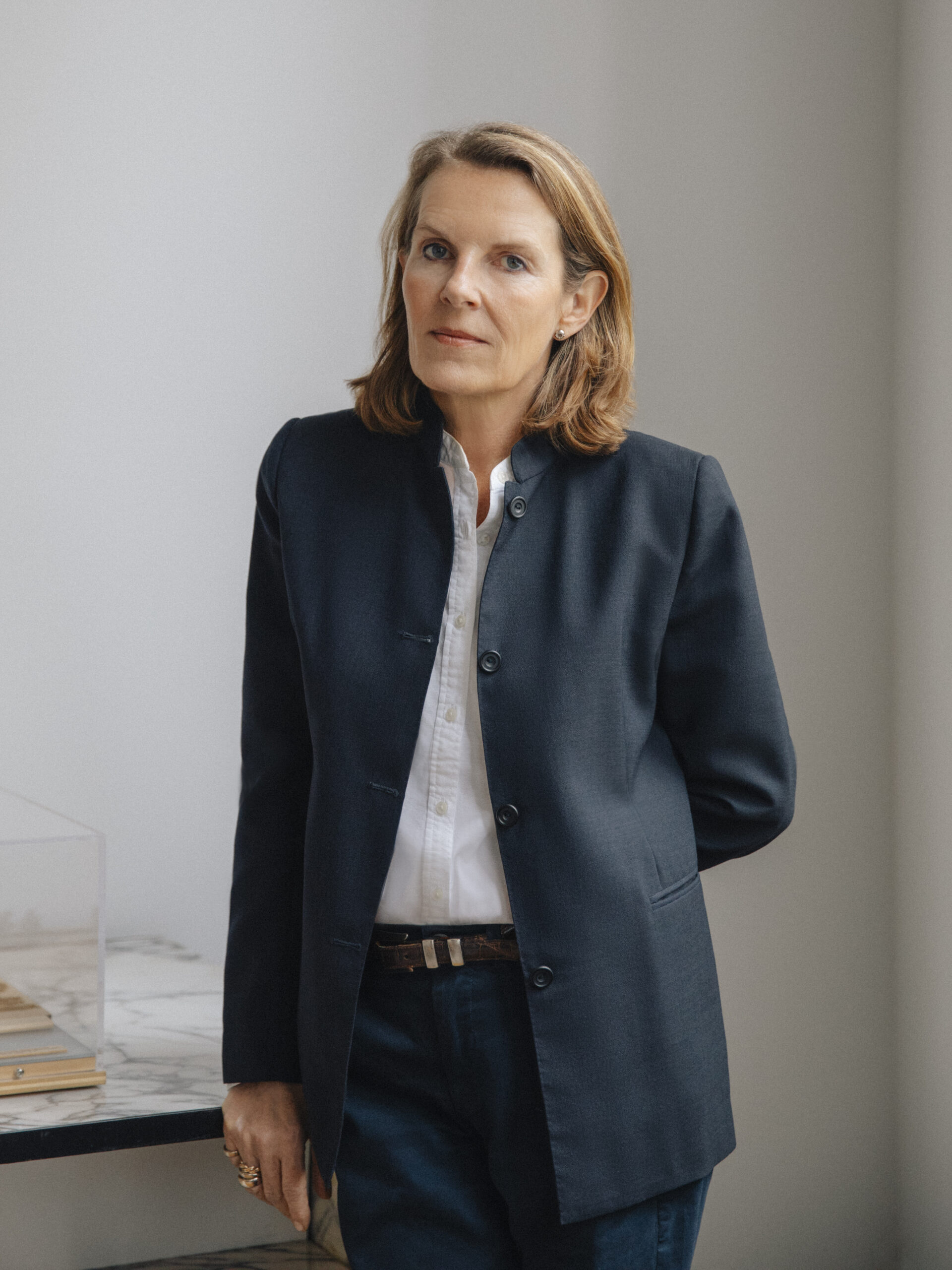 Annabelle Selldorf poses with a neutral expression in a dark blazer and white shirt, standing next to a marble-topped table against a minimalist backdrop.