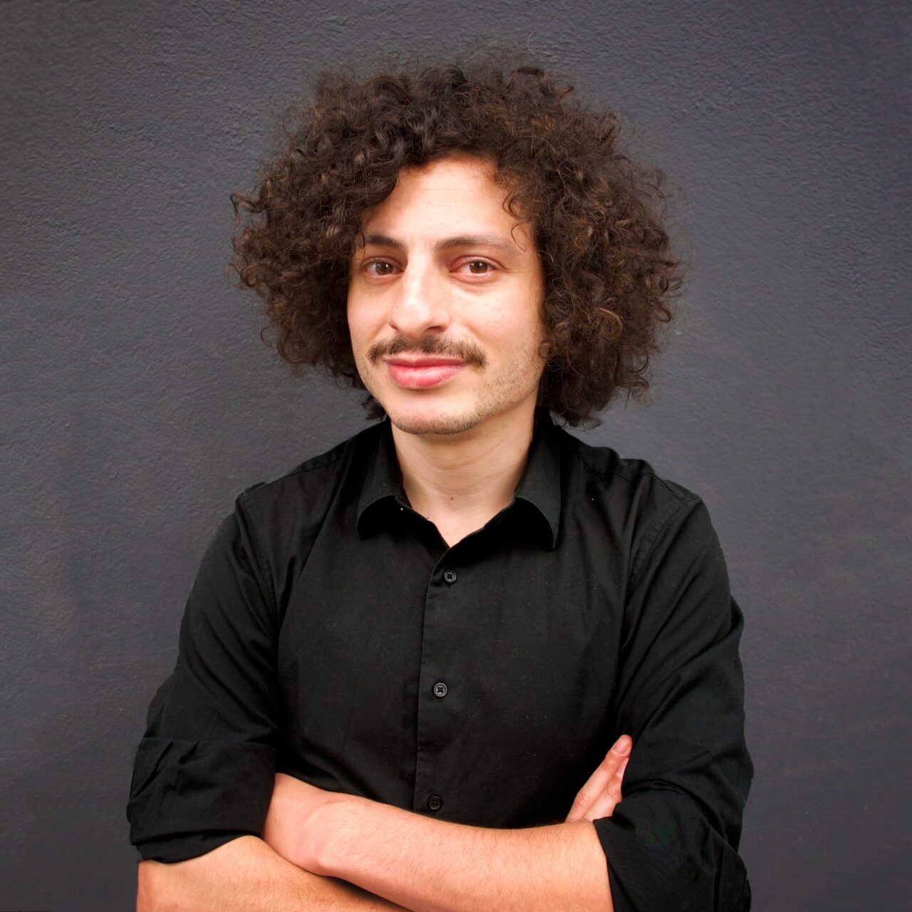 A medium shot of Farzin Lotfi-Jam with curly hair and a mustache, posing with crossed arms in a black button-down shirt against a textured dark gray wall.