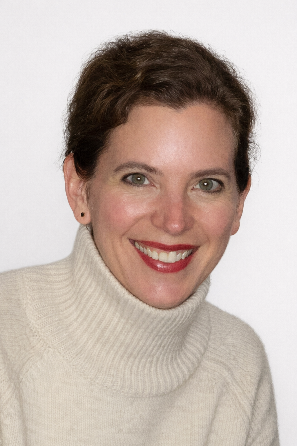 A portrait of a smiling woman with short brown hair and red lipstick, wearing a cream turtleneck sweater against a plain white background.