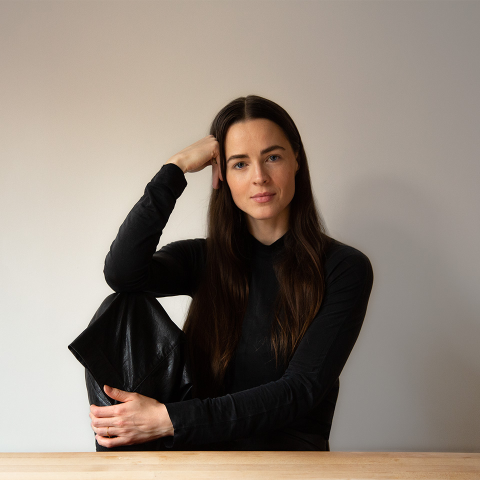 A portrait of a woman with long dark hair wearing a black long-sleeved shirt, resting her head on her hand while sitting at a wooden table against a plain light wall.
